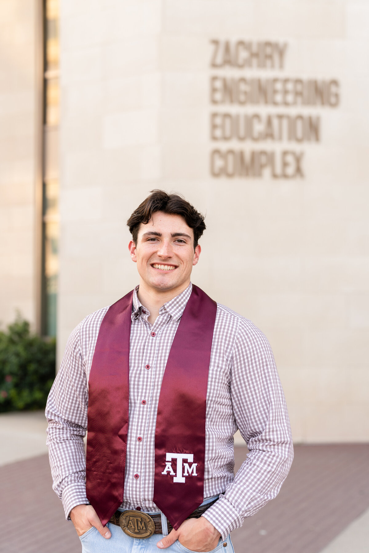 Texas A&M senior guy standing with hands in jeans pocket while wearing a checkered button up and Aggie stole with Zachry Engineering Building behind him