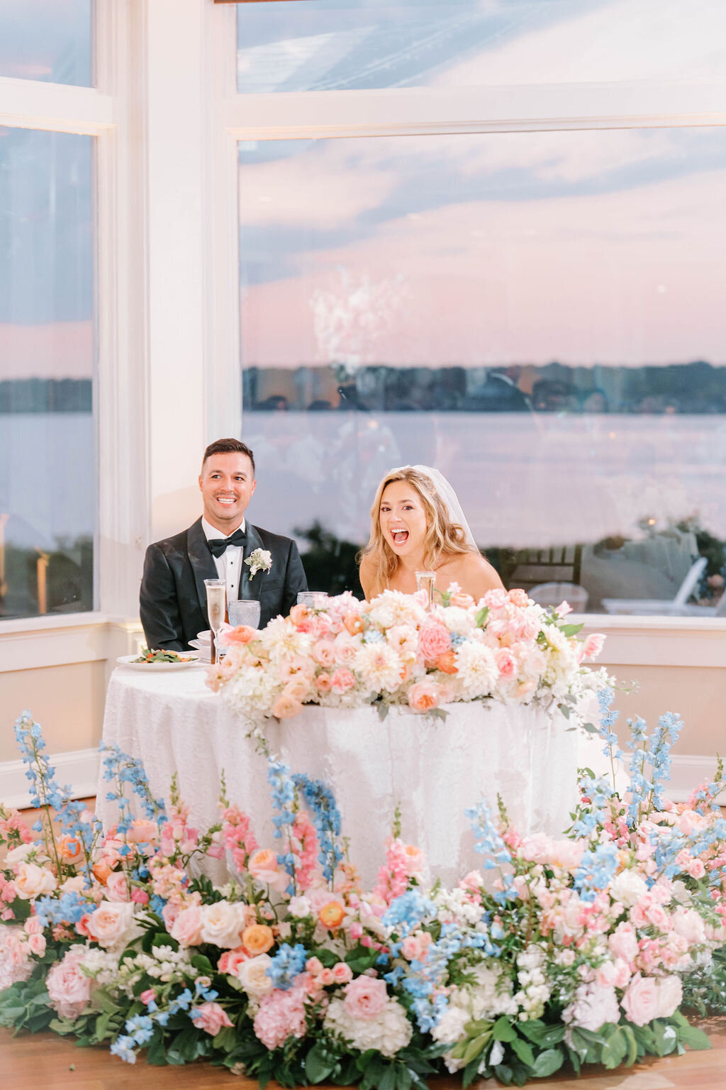 Rhode Island Wedding Photographers | A joyful couple in wedding attire sits behind a flower-adorned table, with a serene lake and pastel sunset visible through a large window.