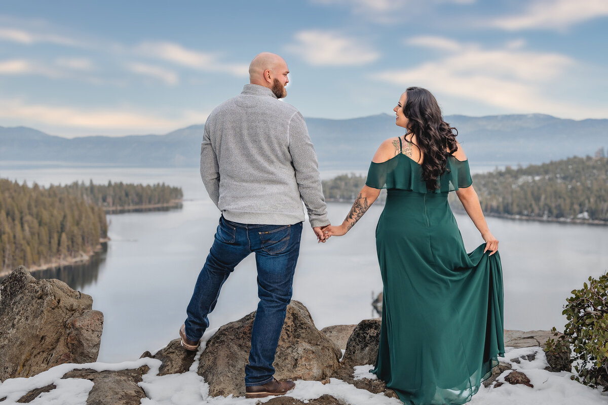 Engaged couple look at each other and hold hands as they overlook lake tahoe's emerald bay.   The girl is wearing a green long dress. Photo by wedding photographer, philippe studio pro.