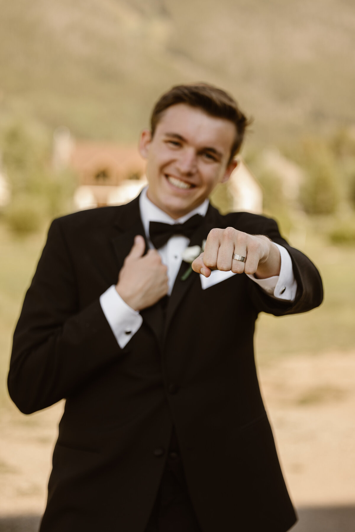 A groom proudly shows off his wedding ring with a playful smile, wearing a classic black tuxedo and standing in a sunlit outdoor setting with trees and a blurred building in the background.