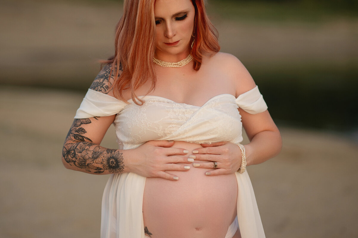 A mother to be stands on a beach holding open her maternity gown to show her bump