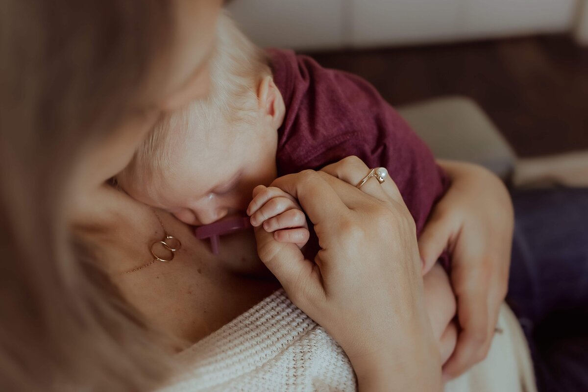 A baby in a maroon outfit sleeps on a woman's chest, holding her hand. The woman wears a white top and a ring with a pearl.