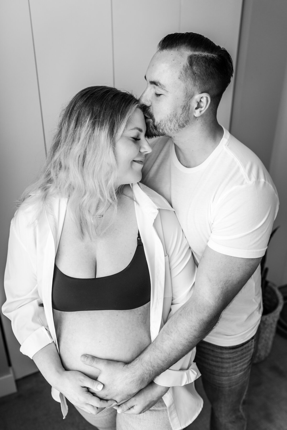 A pregnant woman in a black bra and white shirt is embraced from behind by her partner, who gently kisses her head while both smile contentedly.