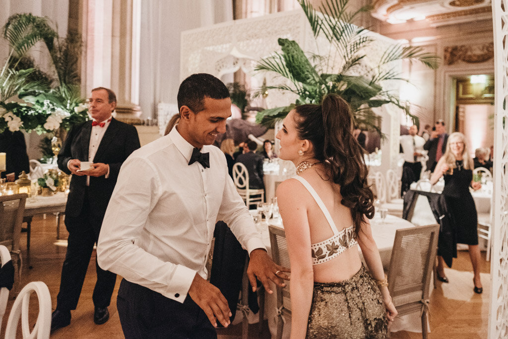 The photo was taken at the Mellon Auditorium in Washington D.C., known as one of the best luxury DC venues with European flair, captured by photographer Sarah Bradshaw. In the image, a handsome man in a white shirt and black bow tie is gracefully paired with a woman in a stunning backless, silver sequin dress as they dance and share smiles at a formal event. The ambiance of the room is exquisitely adorned with white furniture, large green plants, and intricate details, creating a sophisticated and elegant setting. Meanwhile, the background reveals other guests mingling and adding to the lively atmosphere of the occasion.