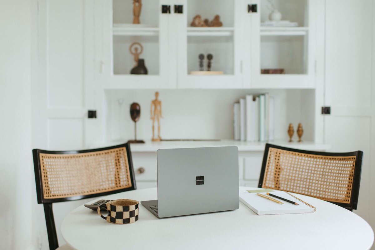 Two empty black and tan rattan chairs behind a table with a laptop, notebook, and checkered mug.