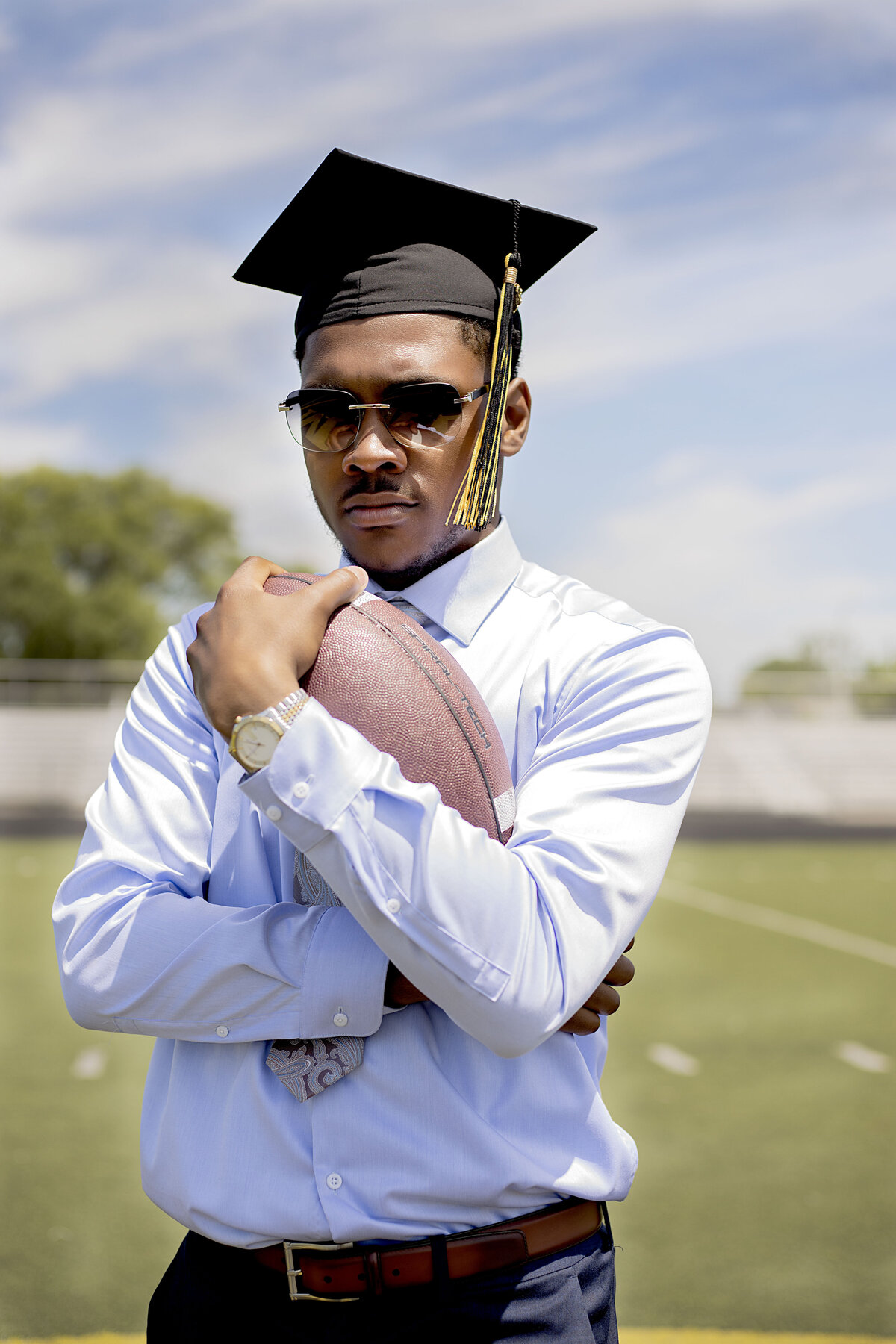 Senior boy with light blue dress shirt, navy blue slacks, sunglasses, and a watch wearing graduation cap holding a football on the football field