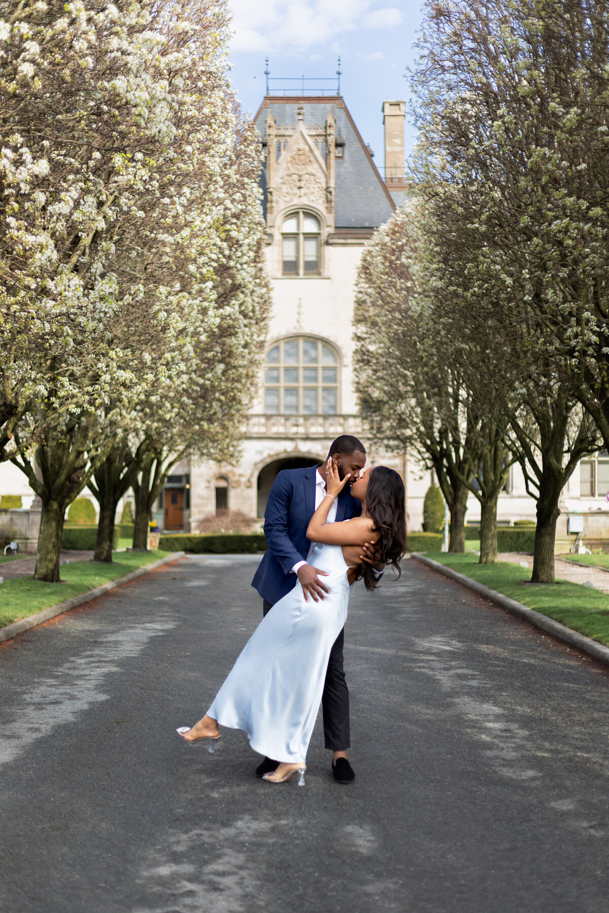 Ochre Court Mansion  | Kelsey Sheehan Photography Timeless Rhode Island Weddings | A couple embraces on a tree-lined path in front of a historic building. The scene is romantic, with blooming trees and an elegant atmosphere.