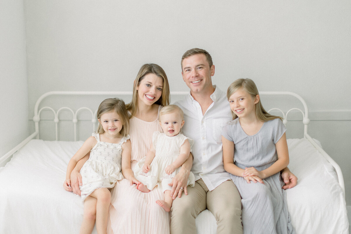 A family portrait of 5 while sitting together on a bed by a window at a photography studio in the Fort Worth area by a luxury family photographer.