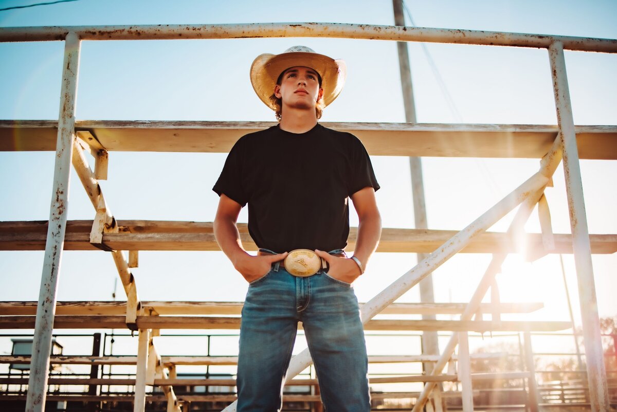 A senior wearing a cowboy hat and large bet buckle posing beneath the bleachers.
