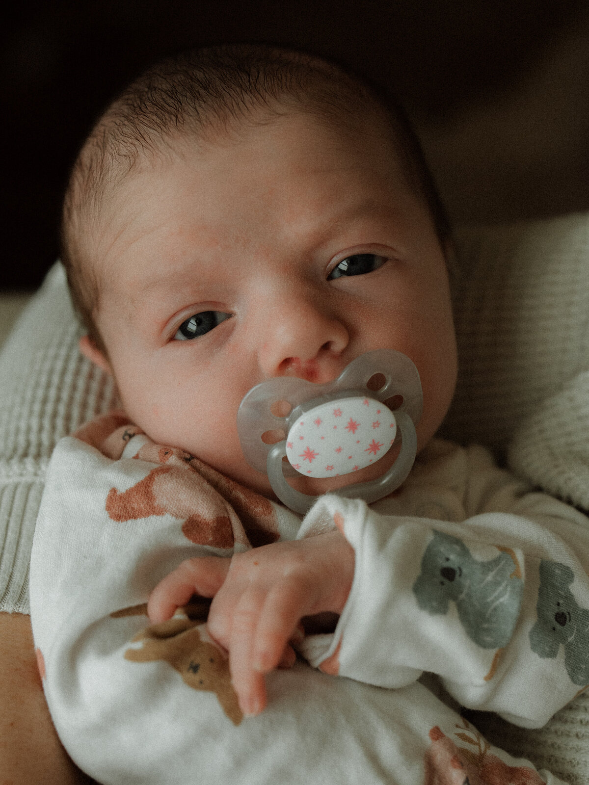 Newborn with bright eyes looks at camera while sucking a pacifier during an at-home newborn photography session.