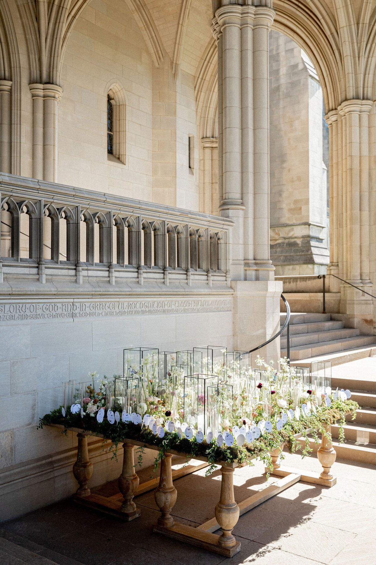 The photo was taken at Washington National Cathedral in Washington D.C. by photographer Twah Dougherty. In the stunning setting of the cathedral, a floral arrangement sits on a wooden cart under an arched stone structure, featuring lush greenery and assorted colorful flowers. The background includes stone walls, intricate carvings, and steps leading to an elevated platform, creating a beautiful and serene atmosphere. Additionally, Current Films can capture the beauty of this setting as talented videographers.
