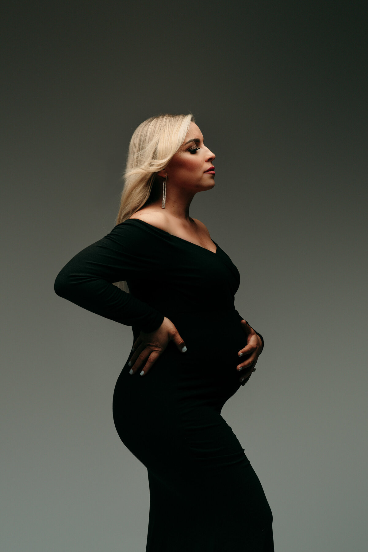 Maternity portrait of pregnant blonde woman wearing fitted long sleeve black dress holding baby bump
