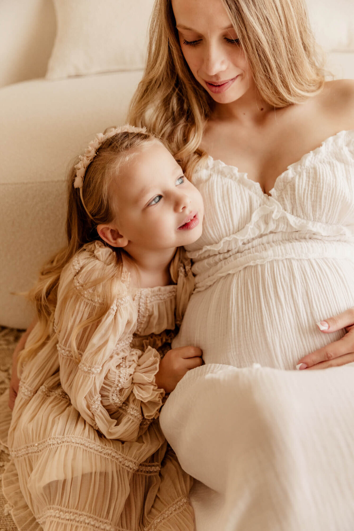 Girl with blue eyes wearing a pink dress looks to the side while sitting next to her pregnant mother.