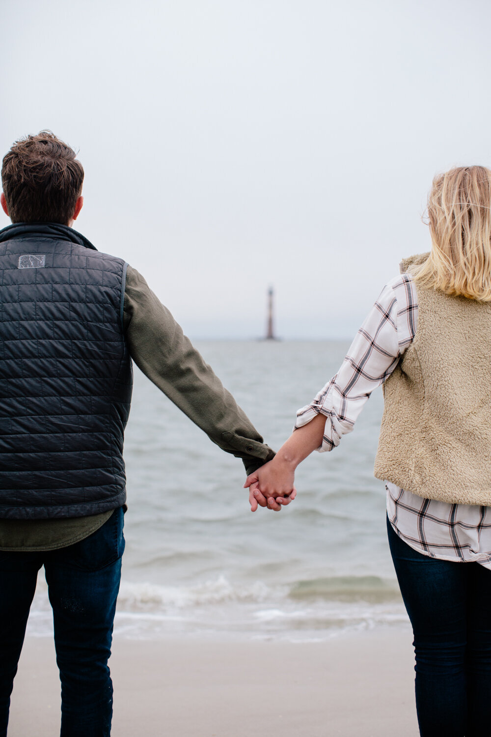 Engaged couple holding hands with  lighthouse in background at Folly Beach SC. Charleston engagement photos photographed by Stephanie Bailey Photography.
