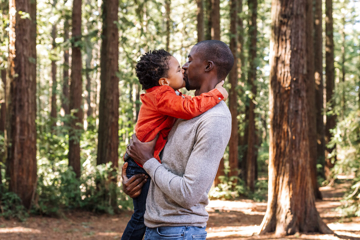Dad holding and kissing  4-year-old son wearing an orange jacket in Oakland woods