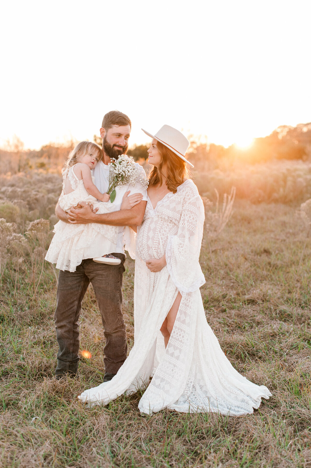 Boho styled maternity photoshoot standing in a tall grass field at golden hour with a beautiful sunset in the background.