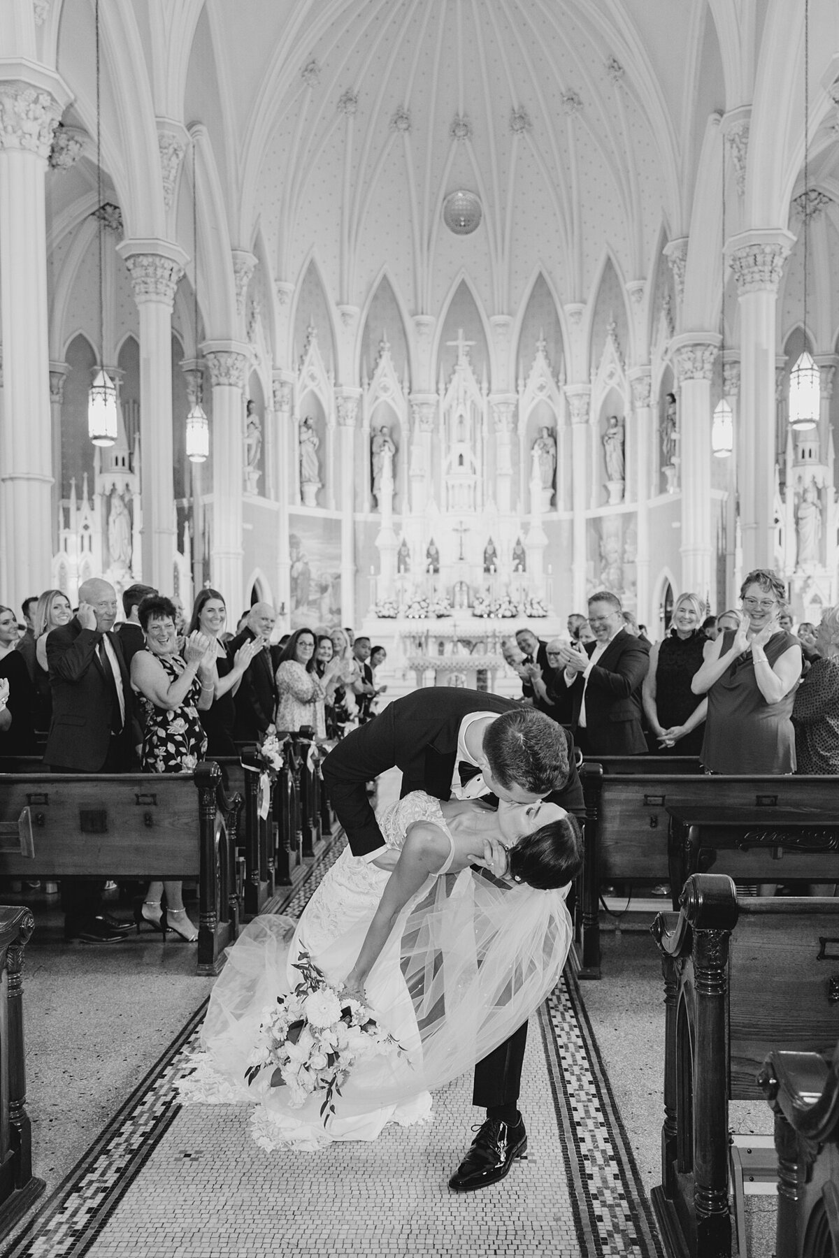 black and white wedding photo in a Catholic church chapel with groom dipping his bride and kissing her passionately at the end of the aisle with the cathedrals ornate architecture behind them