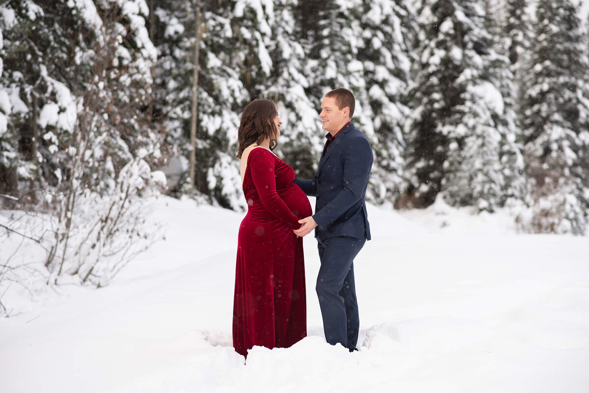 A husband and wife who is pregnant stand looking at eachother with a beautiful winter background. Captured by Brittany Danielle Photography