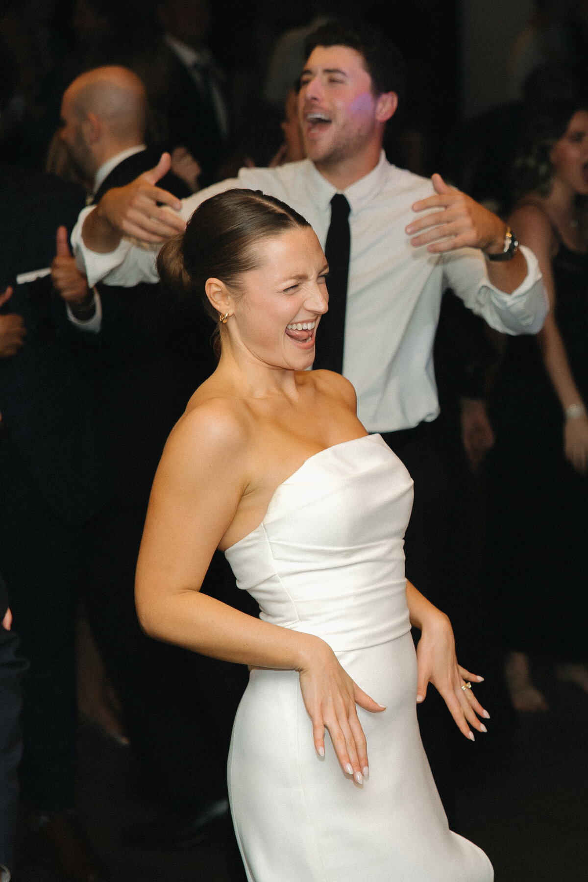 A bride in a strapless, fitted wedding dress flashes an angsty smile with her hands in front of her hip. A man in a white dress shirt with a black tie dances behind her. 