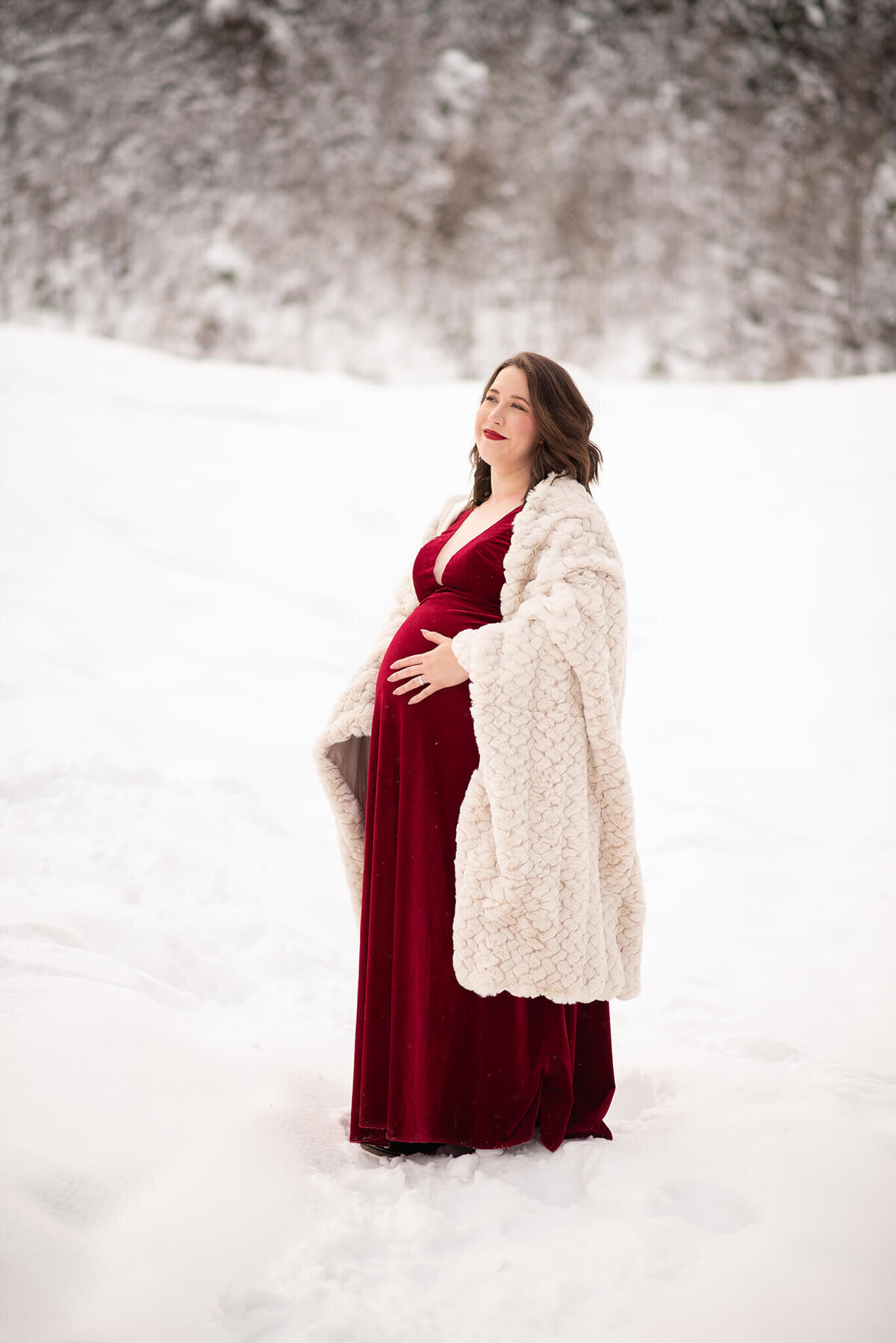 A pregnant woman wears a red dress and stands under a blanket. She is pregnant and stands in the snow. Taken in Sun Peaks by Brittany Danielel