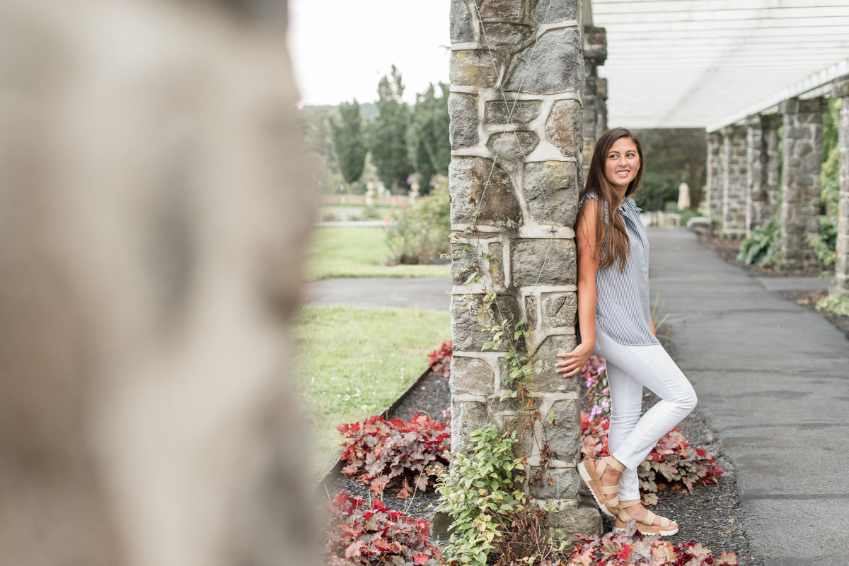 Senior girl smiling at camera leaning back against stone column with right knee bent at Masonic Village in Elizabethtown, Pennsylvania.