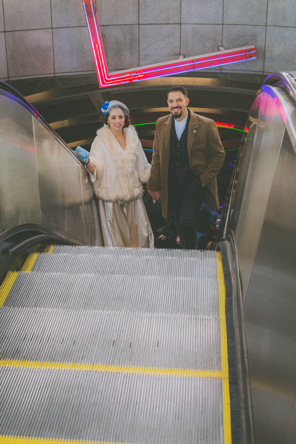 Bride and groom smile while coming up escalator.