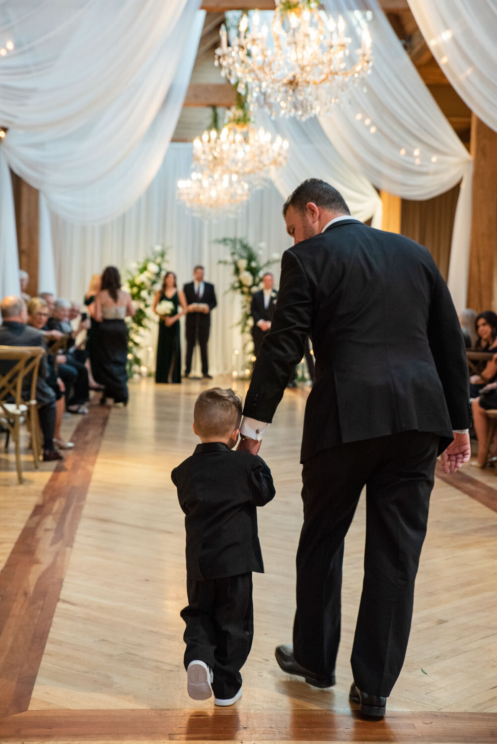 A man in a suit holds the hand of a little boy in a tuxedo