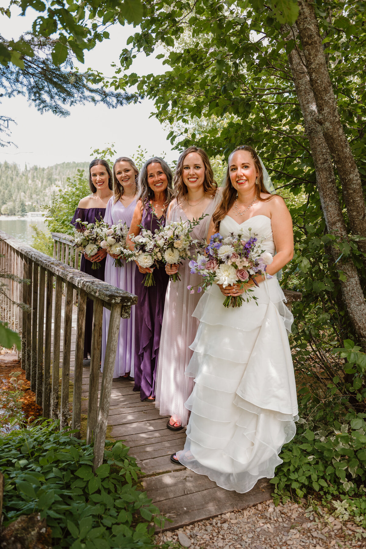 Bride with bridesmaids in lavender and plum dresses holding romantic garden-style bouquets with roses, lisianthus, and larkspur. Lakeside summer wedding in Flathead, Montana with floral design by Indigo & Violet Co. 