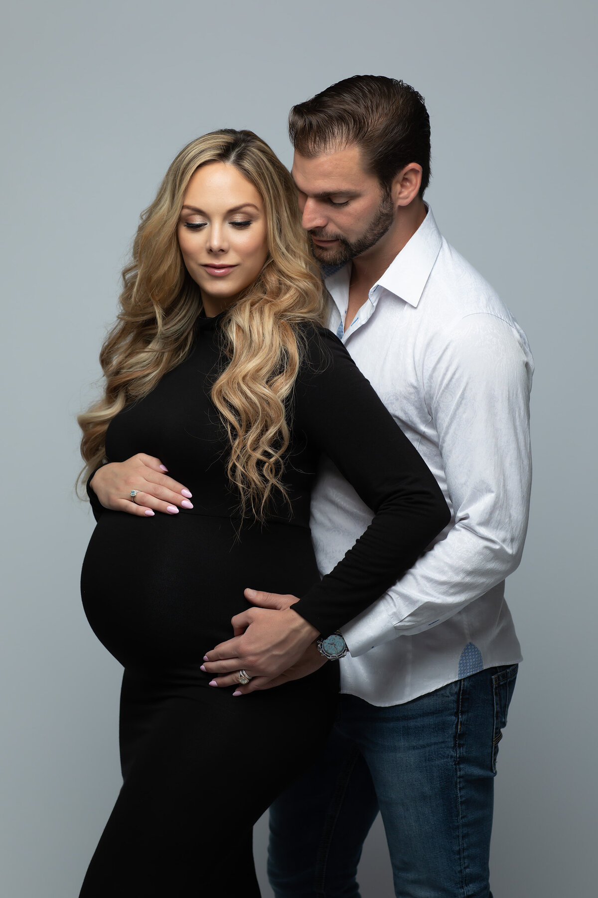 Loving couple posed together with the husband standing behind his beautiful wife, his hand resting on her belly. She is wearing a black fitted dress, has long blonde hair and both are gazing towards her belly.Dallas maternity photography by Lindsay Walden.