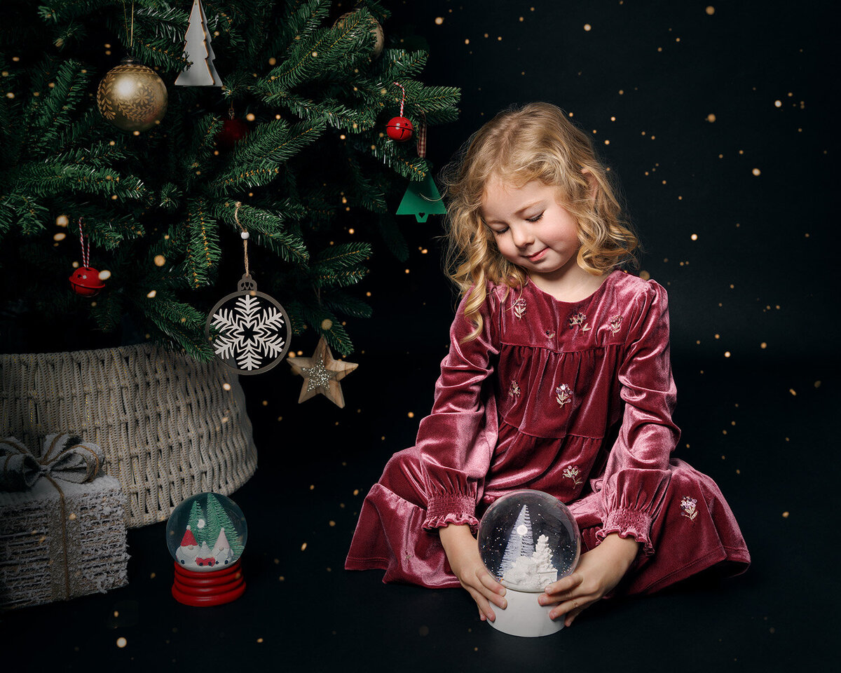Image of a girl looking at a snow globe on a Christmas tree backdrop in the studio. Taken by Norwich portrait photographer Claire Howes. Christmas Mini Sessions Norwich.