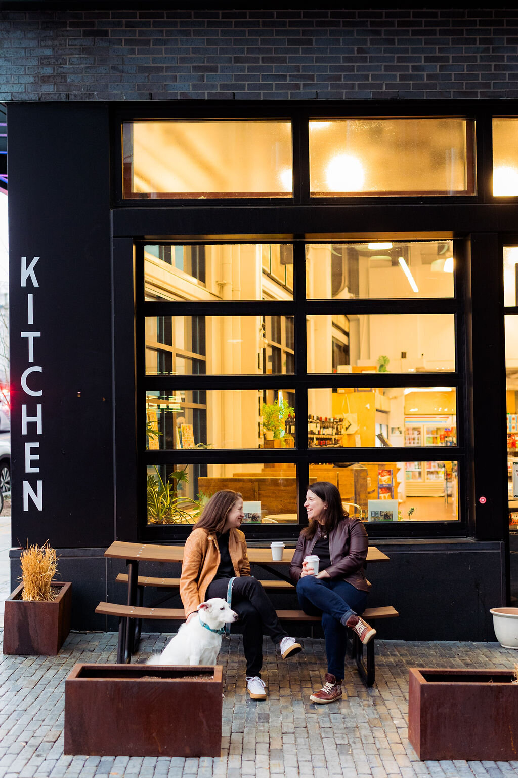 Two people sitting at a picnic table outside a cafe with a dog in front of them