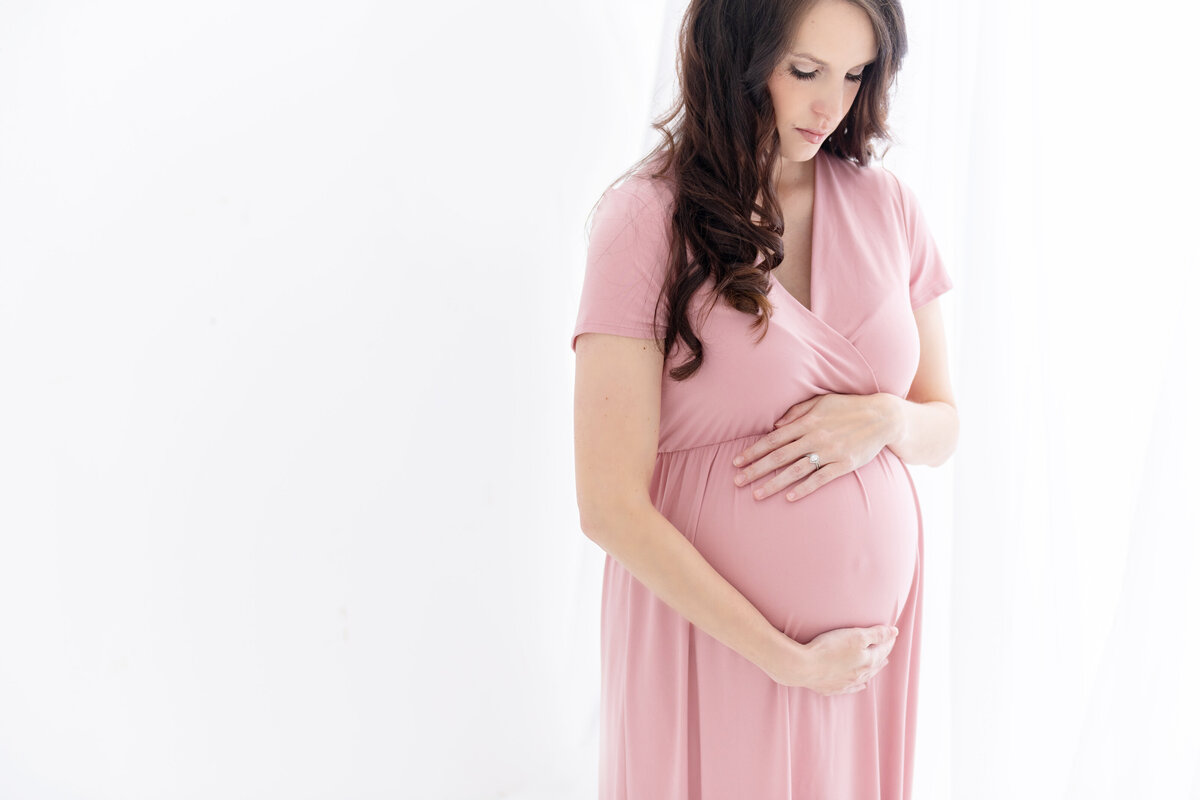 A mother to be holds her bump while standing in a studio looking down in a long pink maternity gown