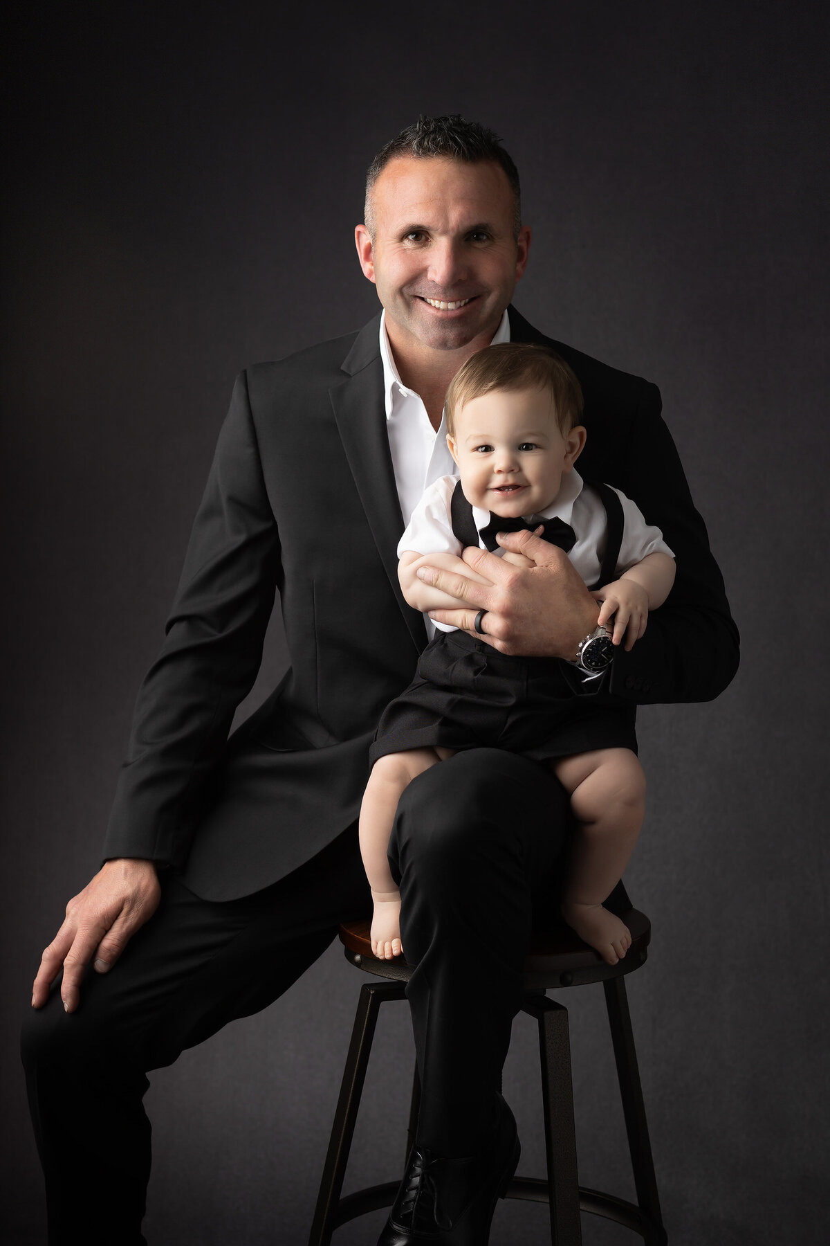 DFW milestone photography featuring a dad in a black suit holding his baby boy dressed in suspenders and a bow tie, seated together in a dark studio backdrop.