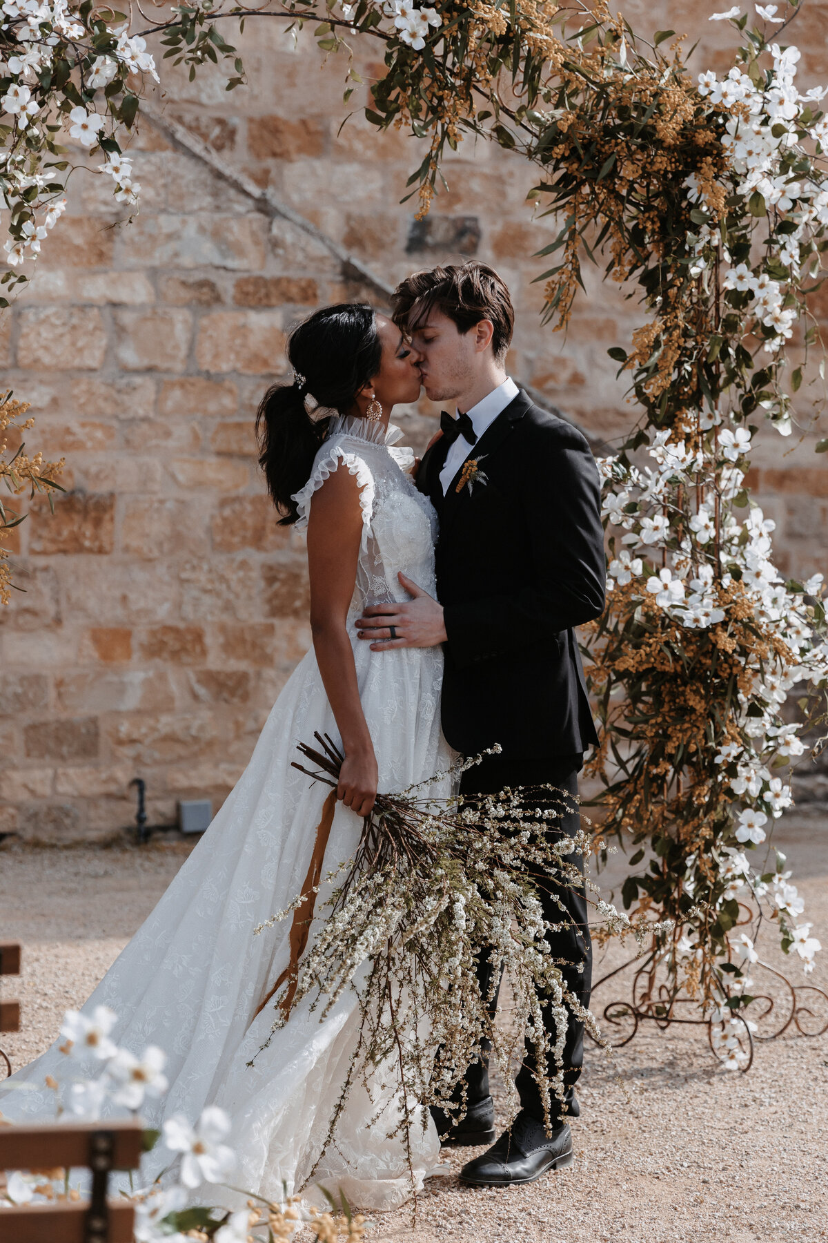 Bride holding a bouquet of wildflowers embraces the groom under a floral arch at Sunstone Winery's outdoor ceremony space.