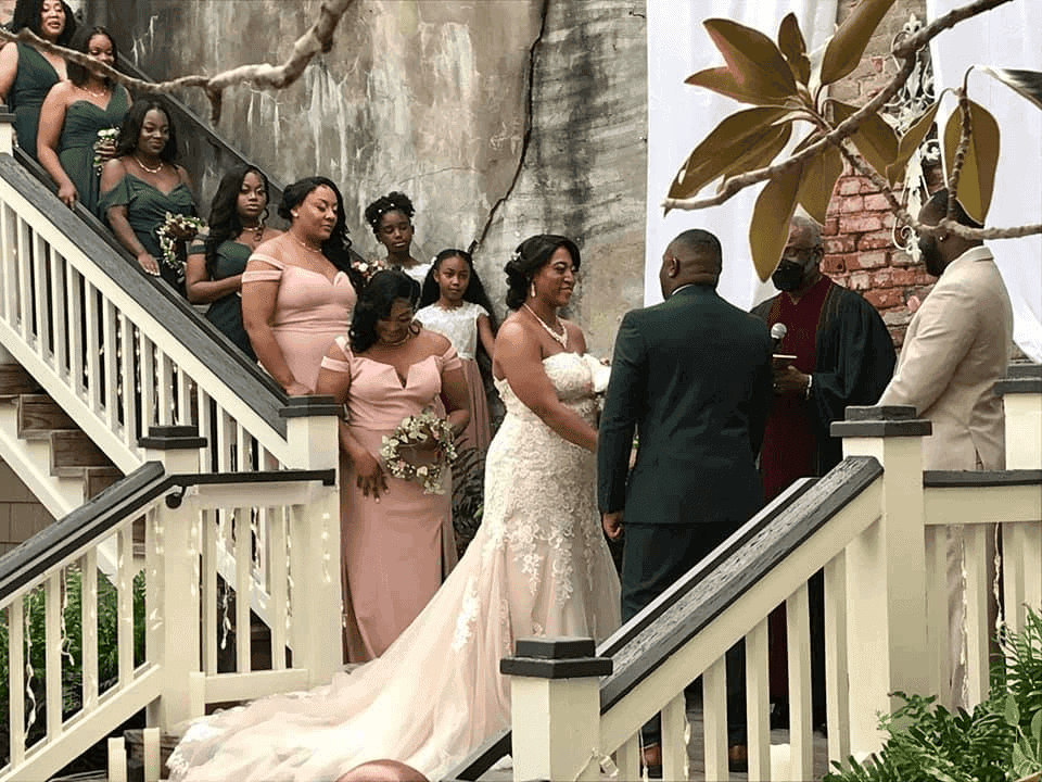 A bride and groom stand facing each other during their wedding ceremony on an outdoor staircase, surrounded by bridesmaids in pink dresses and groomsmen, with an officiant and lush greenery nearby.
