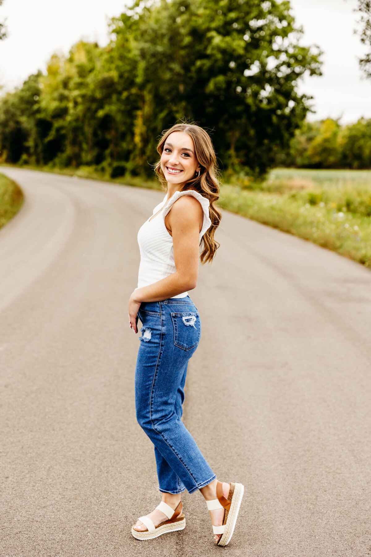 pretty brown haired teenage girl in a white top and jeans standing sideways on a back road and smiling as she enjoys her senior photography session in Neenah.