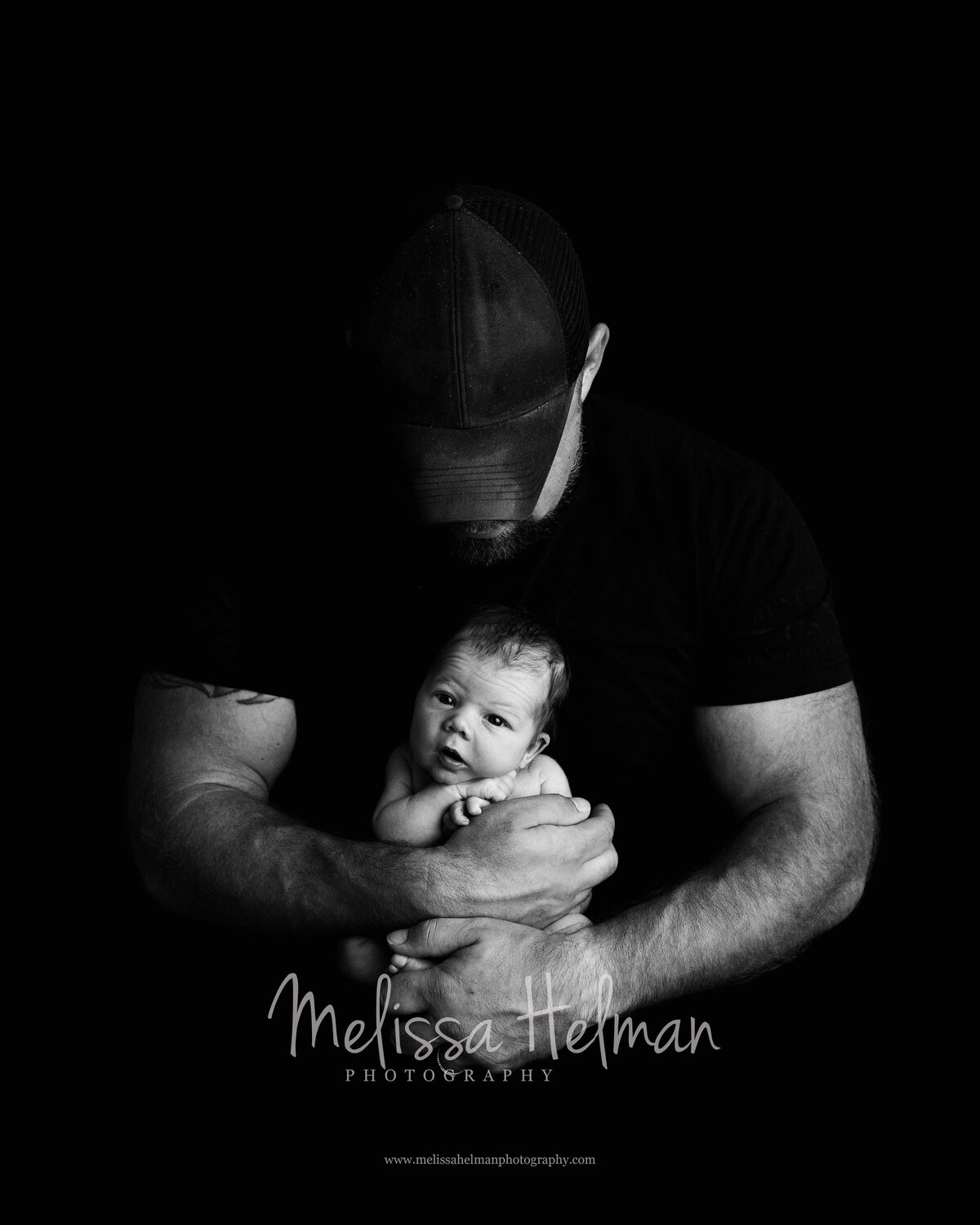 Dad holding baby boy in his hands with his head looking down wearing a hat inPittsburgh photography studio.