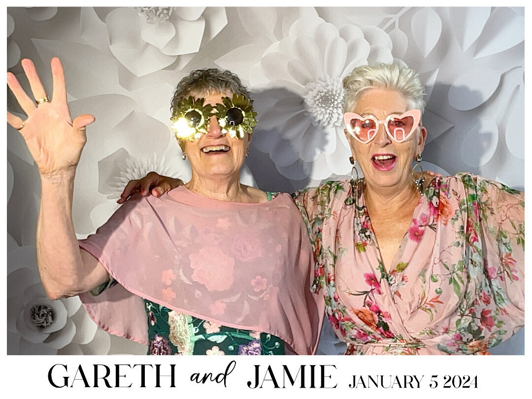 two women in pink in front of a white paper flowers backdrop with fun sunglasses on in a photo booth capture from a wedding at pemberton