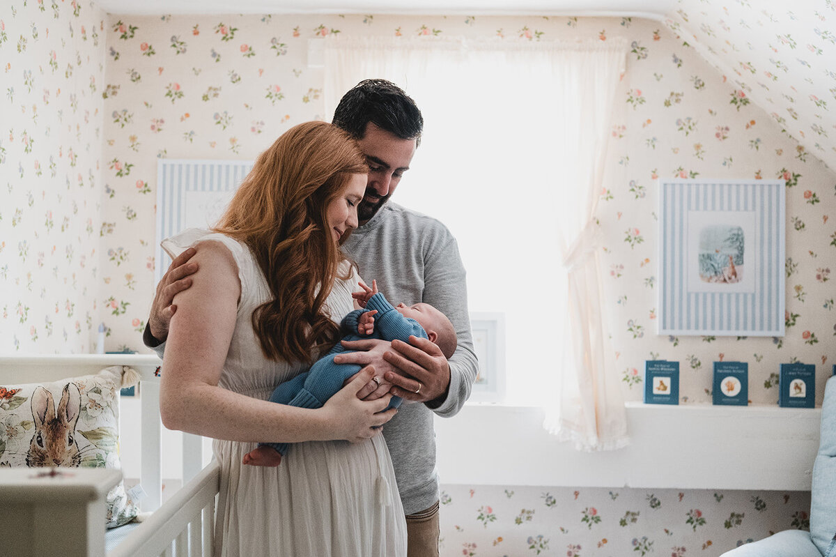 Parents holding their newborn in a bright, cozy nursery with floral wallpaper, captured in a natural, documentary style by a Connecticut newborn photographer