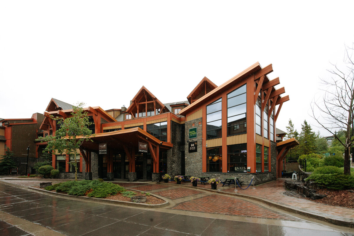 Contemporary timber-and-glass exterior of Mainspace in Canmore, Alberta, featuring striking architectural beams, stone accents, and large windows with views of the surrounding Canadian Rockies.