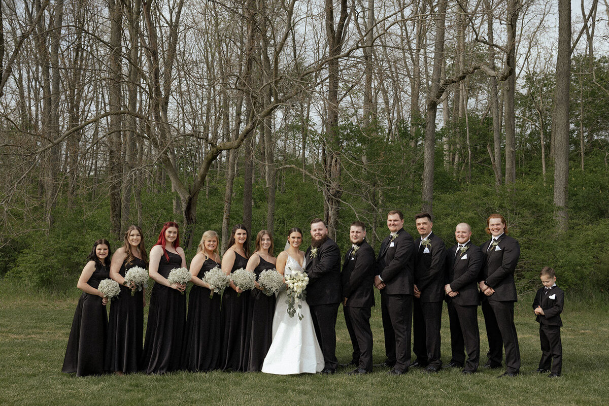 Large wedding party dressed in black and white posing in a wooded area