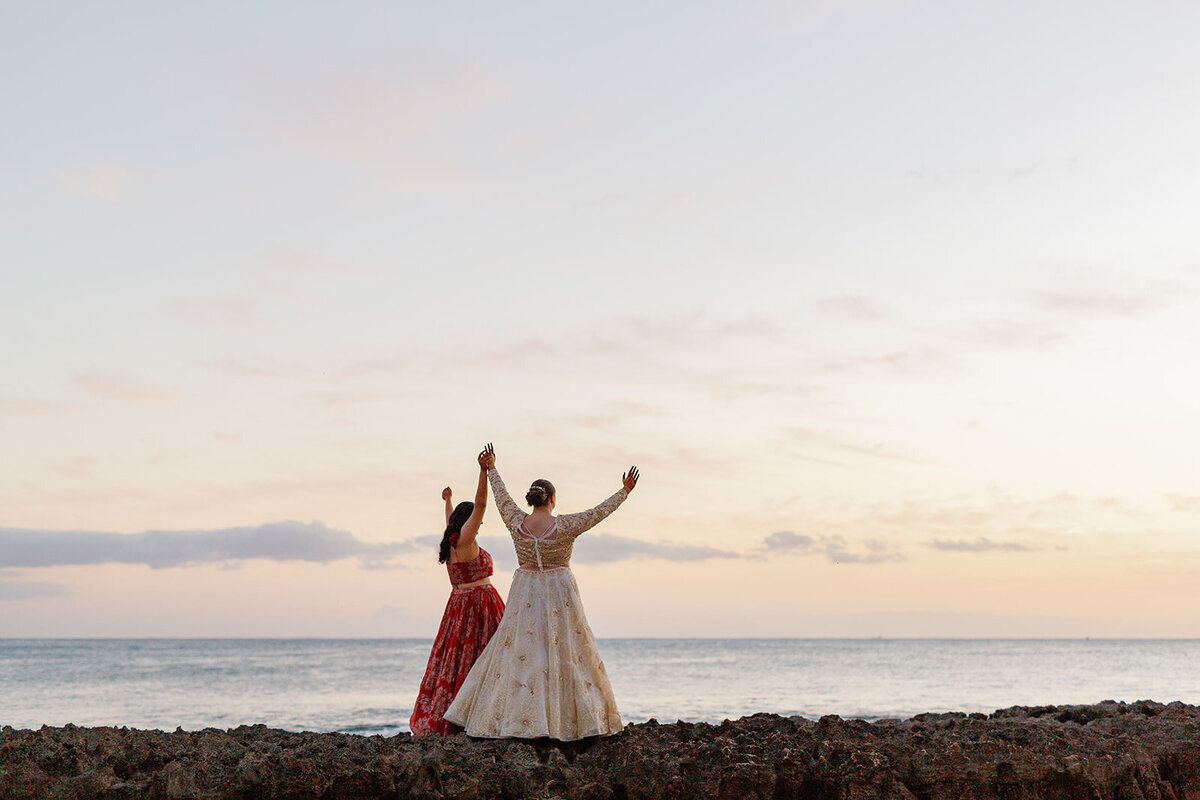 A newlywed couple standing at the edge of the water with their arms up 