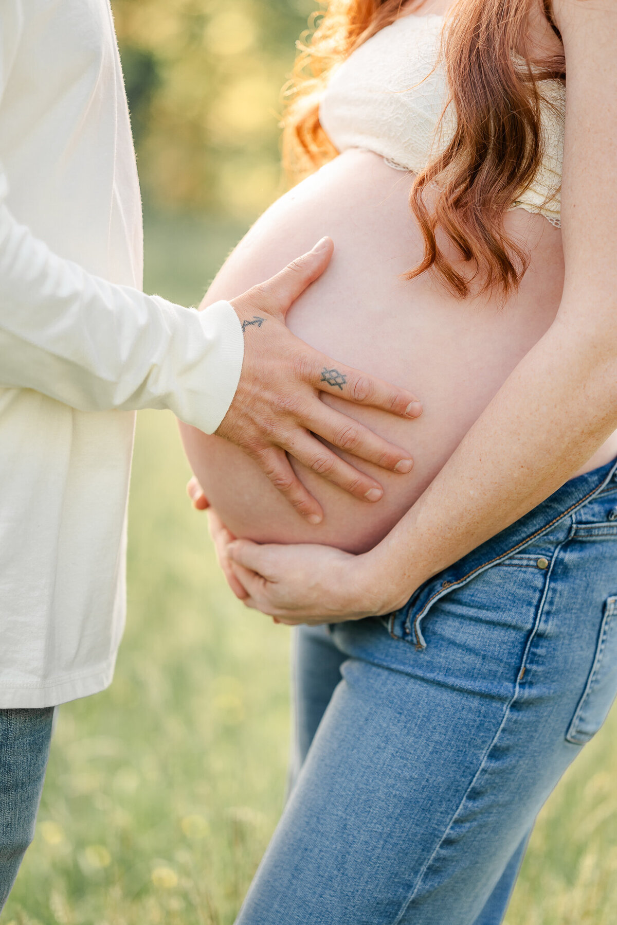 A picture of a pregnant woman in jeans and a white bralette. She is holding her exposed belly while her partner also rests his hand there.