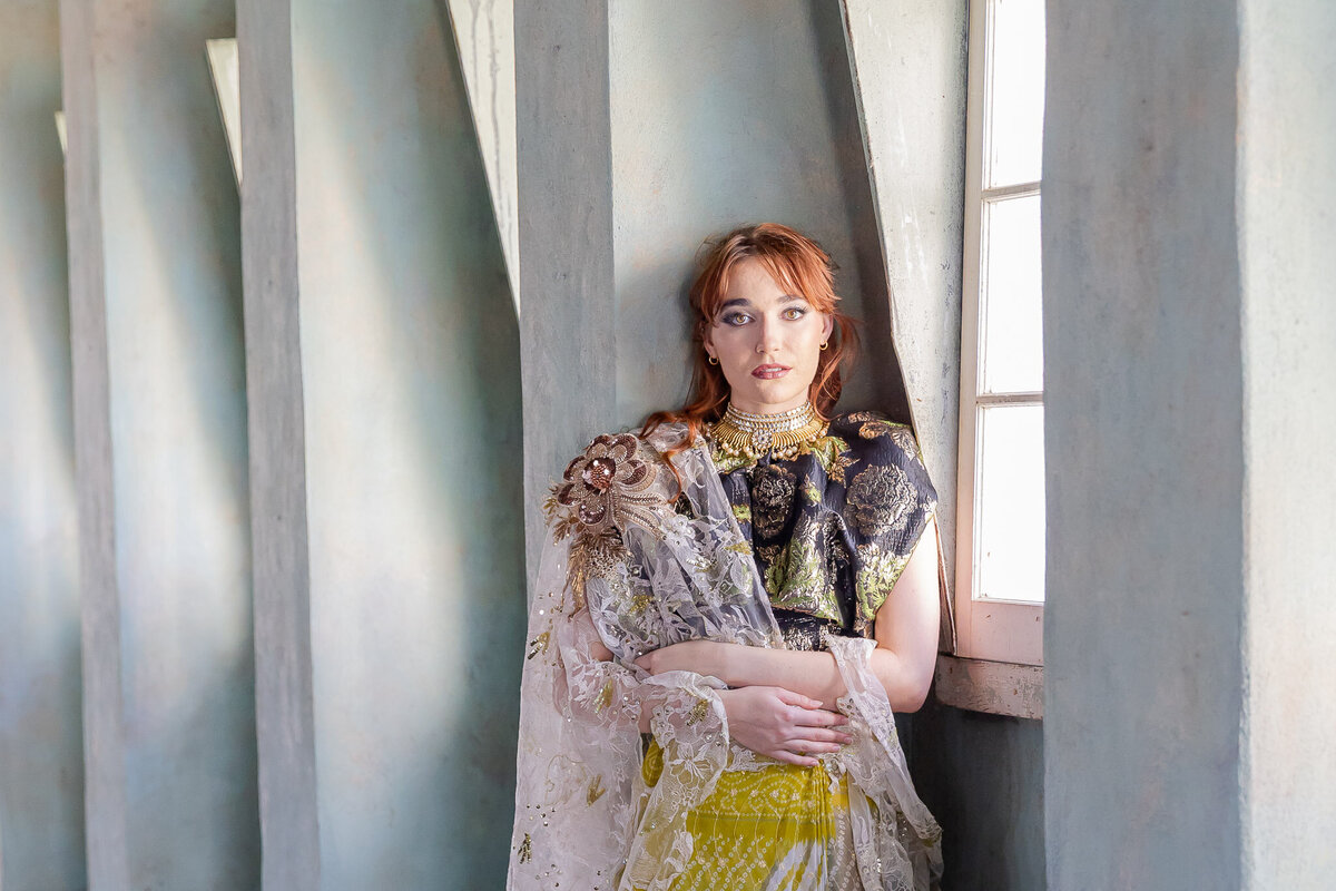 Leaning against a concrete structure, a woman with bright red hair looks at the camera. Her outfit is beautifully eccentric.
