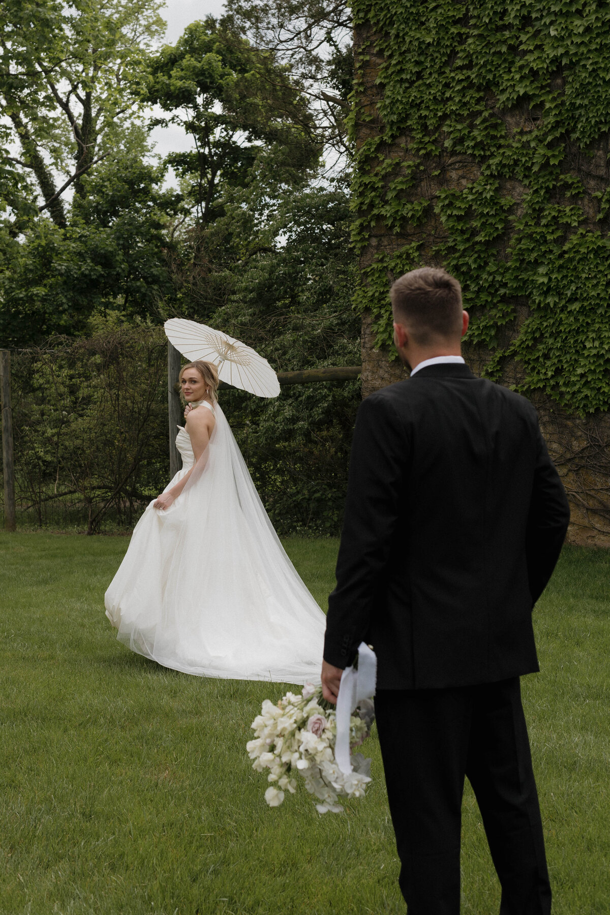 A cinematic storytelling shot capturing a beautiful first look moment between a bride and groom. The bride elegantly holds a white parasol, glancing lovingly at her groom, in a lush, green garden setting. Captured by a documentary-style luxury wedding photographer.