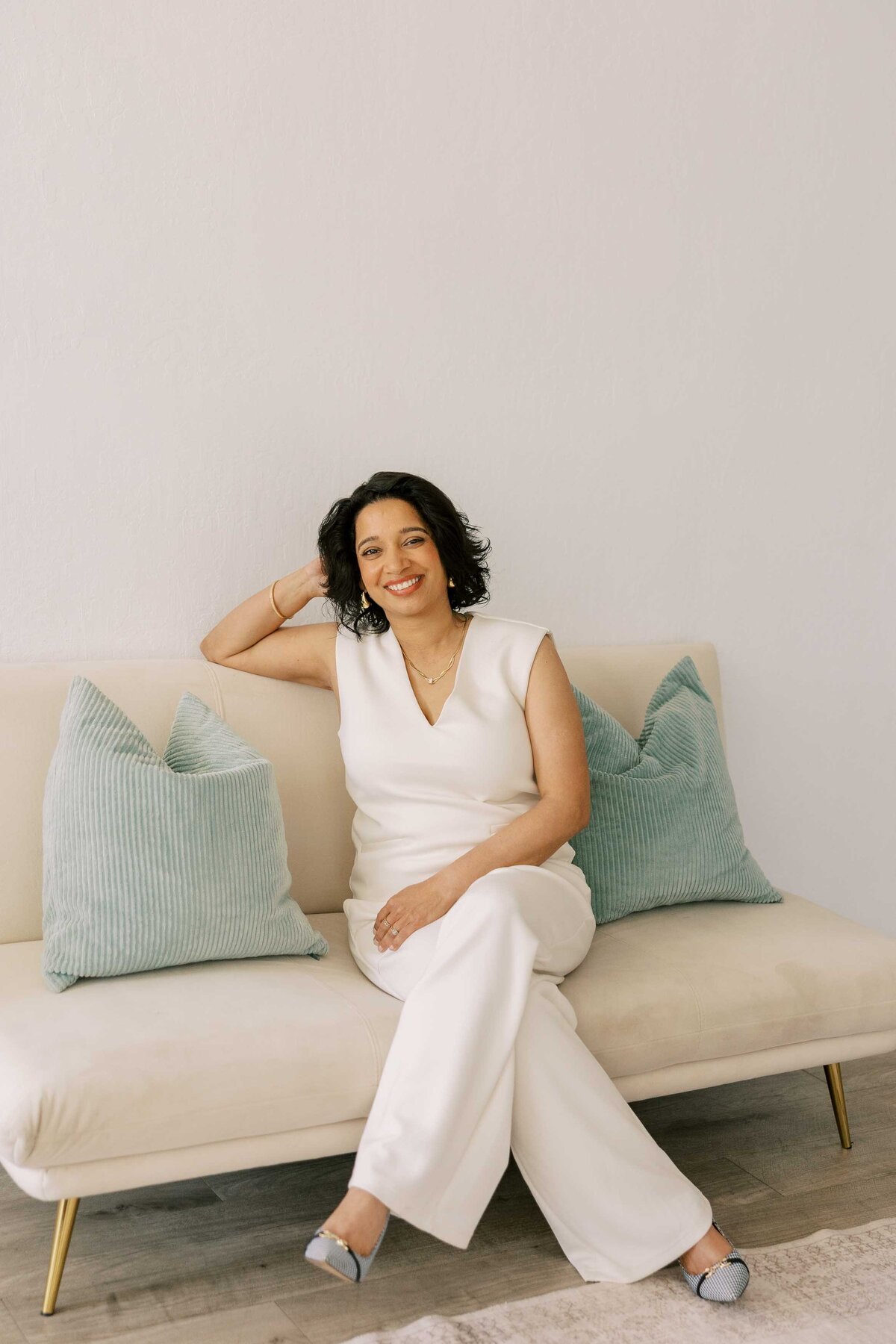 Woman wearing an all-white outfit and leaning on her elbow on a couch with blue pillows.