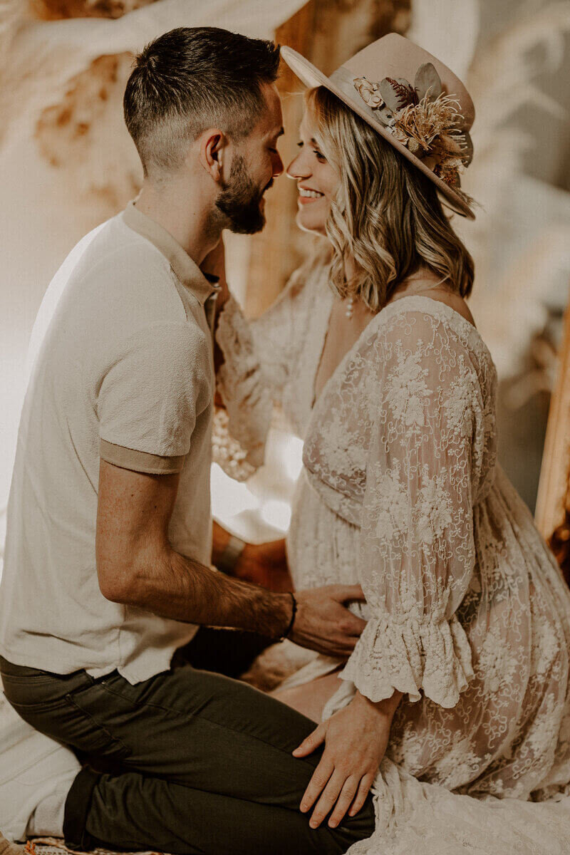 Couple assis face à face, souriant. L'homme caresse le ventre rond de sa femme dans un décor cosy lors d'une séance photo grossesse.