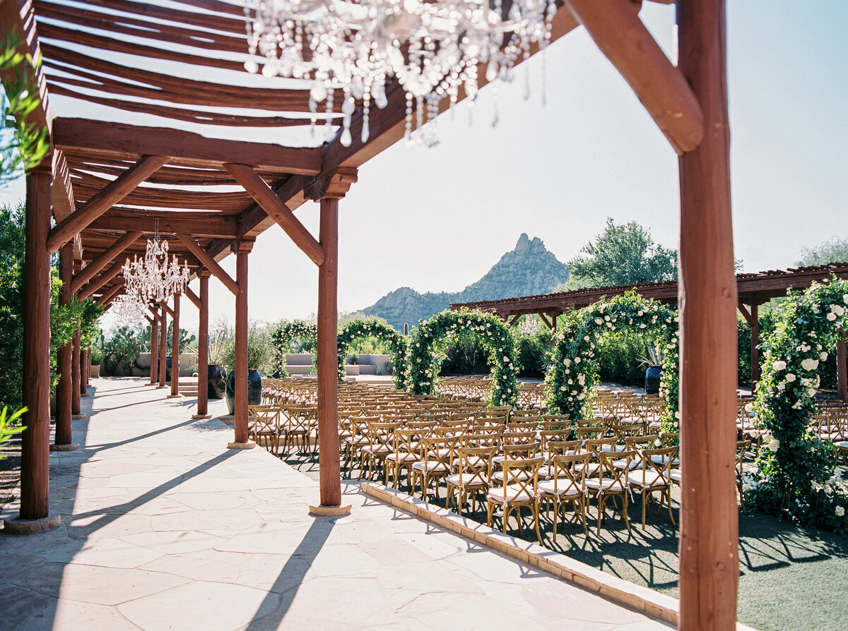 An elegant wedding ceremony setup with floral archways, wooden chairs, and a stunning desert mountain backdrop. A breathtaking location for an outdoor luxury wedding.