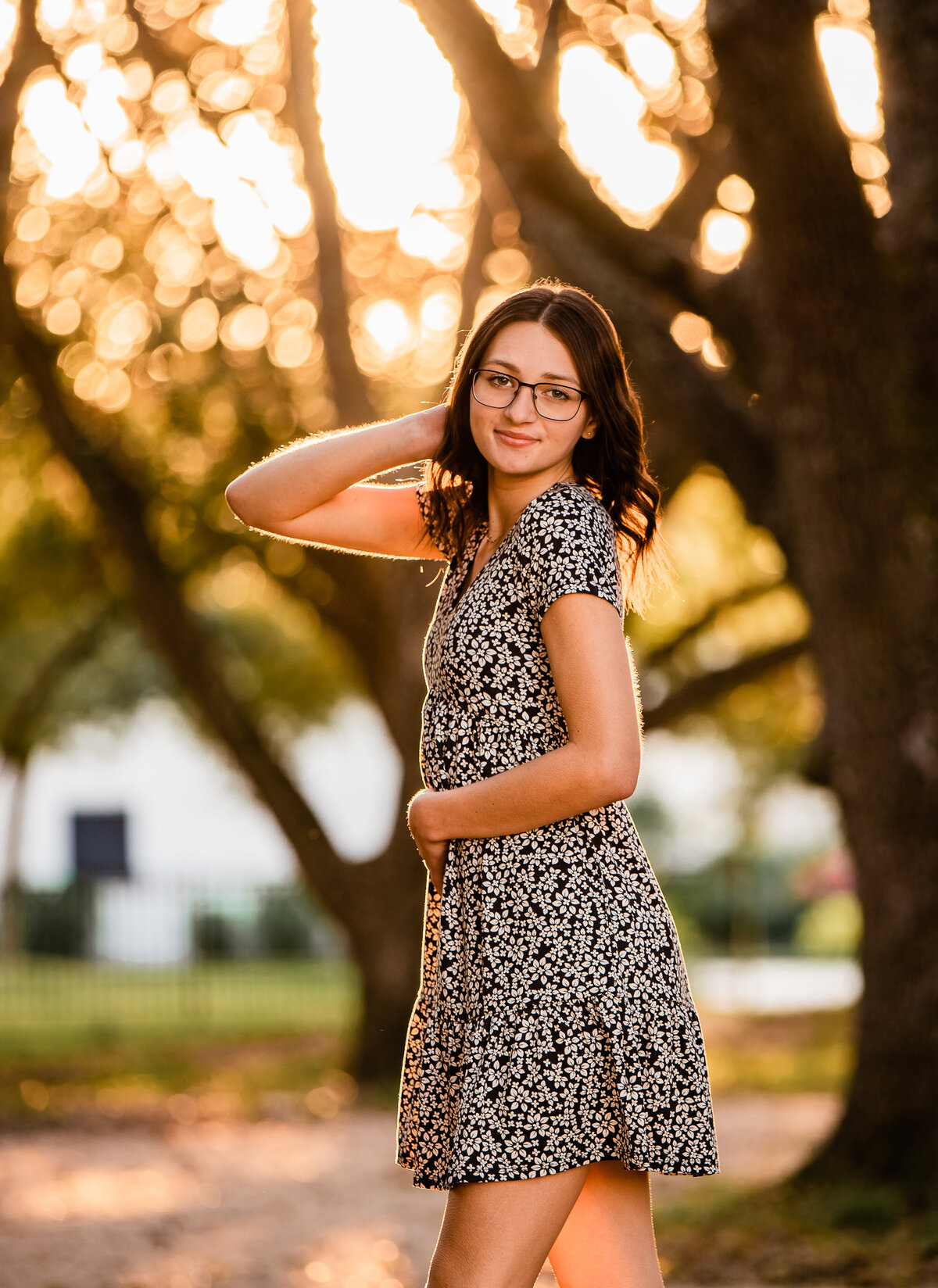 A Houston area graduate stands under a canopy of trees with sunset shining behind her.
