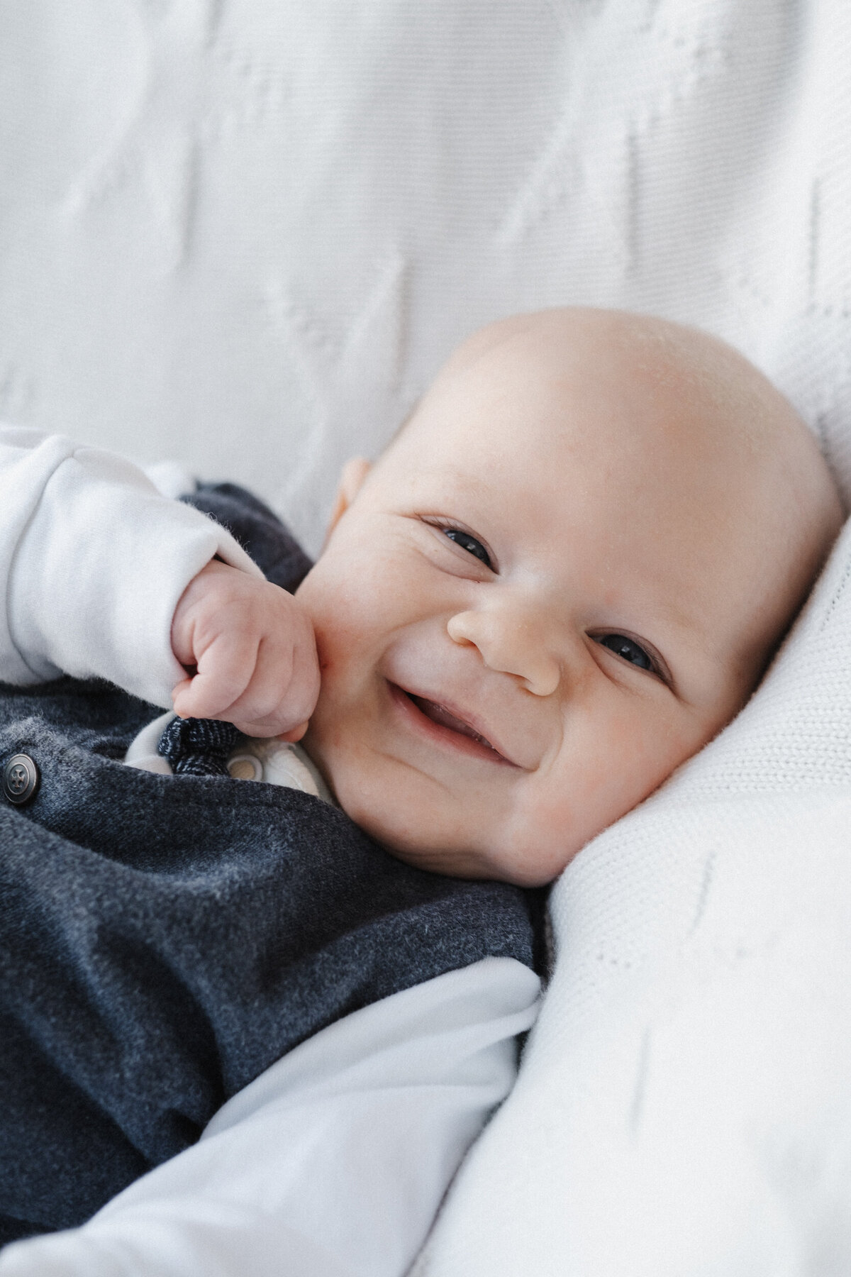 Grinning baby holds fist to his face while smiling and laying on white blanket